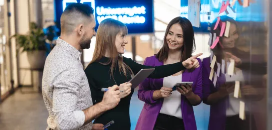Studia II stopnia w Gdyni | Uniwersytet WSB Merito Gdynia: Zdjęcie przedstawiające dwie studentki i studenta pracującego w grupie. Stoją oni przy karteczkach i coś omawiają. Ubrani są biznesowo. Z tyłu widać neon Uniwersytetu WSB Merito