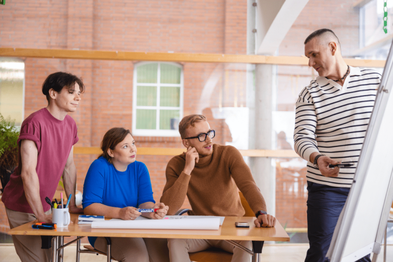 Pedagogika resocjalizacyjna z profilaktyką społeczną | Studia I stopnia w Gdyni | Uniwersytet WSB Merito Gdynia: Trzech studentów przy stole słucha wykładowcy, który tłumaczy coś przy flipcharcie