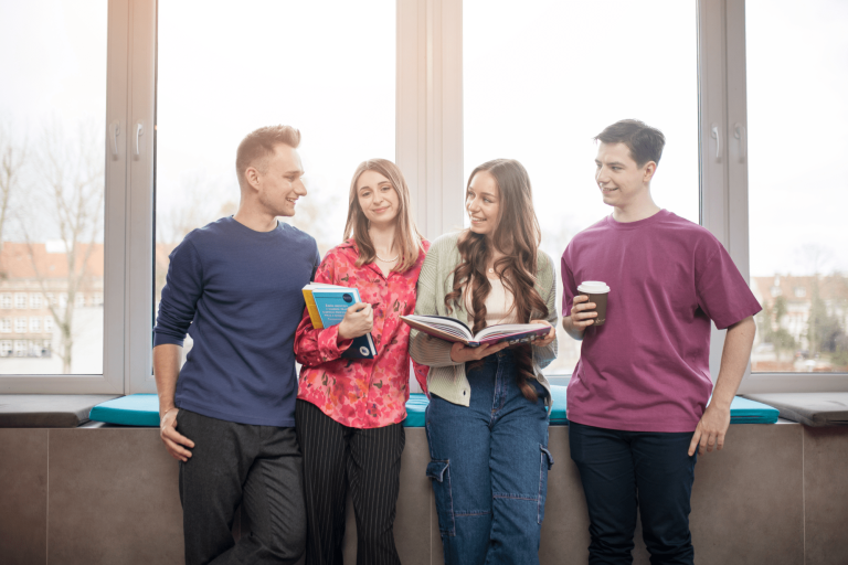 Poznaj nas | Uniwersytet WSB Merito Gdynia: Zdjęcie studentów opierających się o parapet, z tyłu świeci słońce. Na zdjęciu znajduje się dwóch chłopaków i dwie dziewczyny, które zajmują centralne miejsce i trzymają książki
