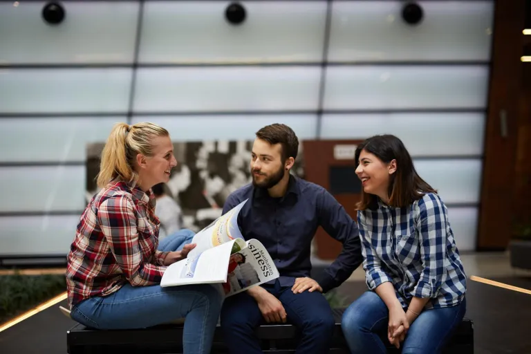 Program firma | Uniwersytet WSB Merito Gdynia: Zdjęcie trójki studentów w tym dwóch kobiet. Znajdują się oni w przestrzeni Europejskiego Centrum Solidarności. Siedzą i rozmawiają