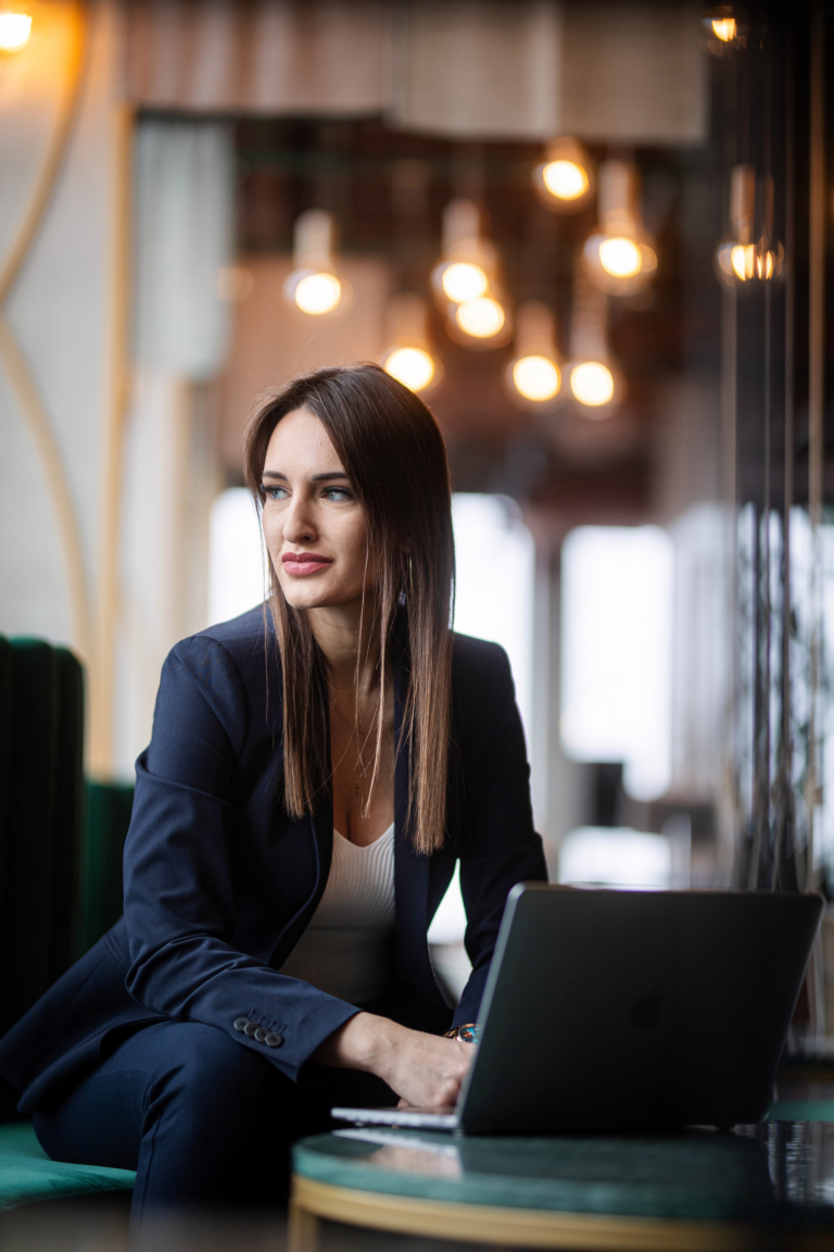 Rankingi i akredytacje | Uniwersytet WSB Merito Gdynia: Zdjęcie biznesowe kobiety o długich blond ciemnych włosach znajdującej się w przestrzeni kawiarni. Jest ona ubrana biznesowo i pracuje przy laptopie. Patrzy w bok