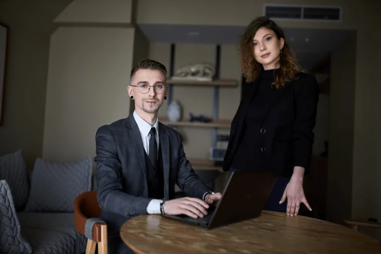 Neuroprzywództwo | Studia podyplomowe w Gdyni | Uniwersytet WSB Merito Gdynia: Zdjęcie osób w średnim wieku. Mężczyzna w garniturze siedzi przy laptopie, obok niego stoi kobieta ubrana na czarno