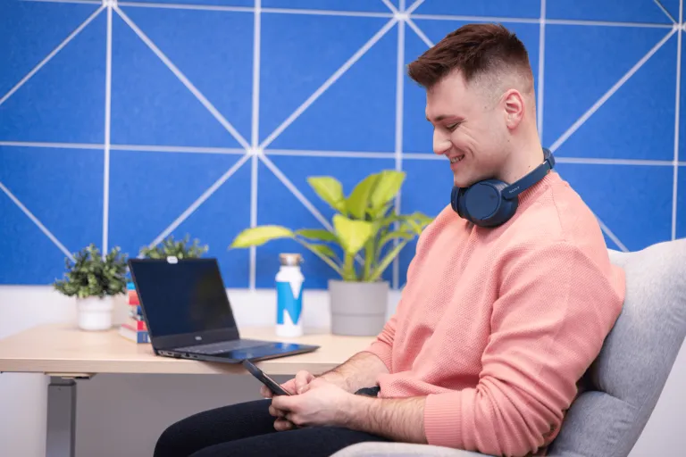 Cyfrowy uniwersytet | Uniwersytet WSB Merito Gdynia: Zdjęcie studenta siedzącego na fotelu i patrzącego w komórkę. Uśmiecha się i ma na sobie brzoskwiniowy sweter. Z tyłu znajduje się ściana w fototapetę w kształcie niebieskim rombów 