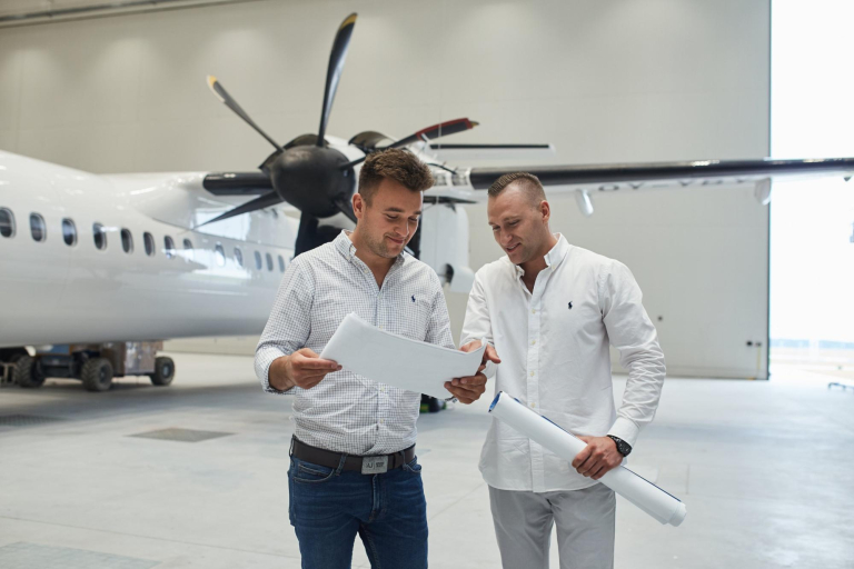 Szkoły | Uniwersytet WSB Merito Gdynia: Dwóch ubranych na jasno mężczyzn patrzy na kartkę i znajdują się w przestrzeni, gdzie jest samolot