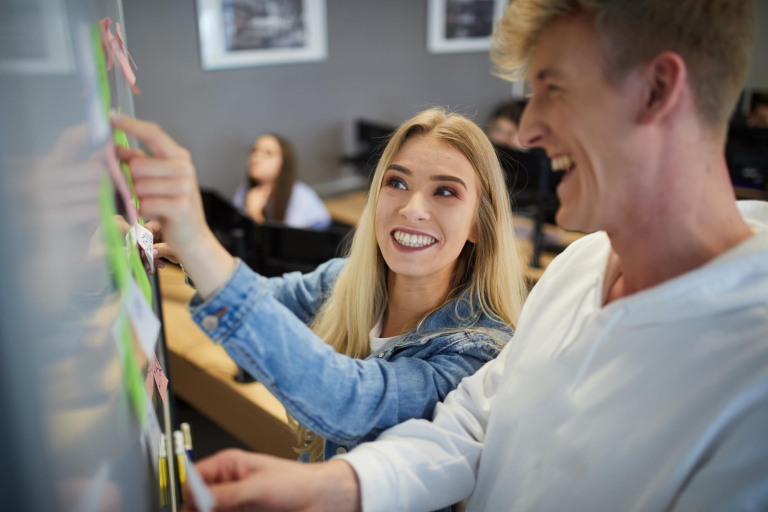 Służby specjalne i policyjne | Studia I stopnia w Gdyni | Uniwersytet WSB Merito Gdynia: Młodzi studenci - on i ona o blond włosach pracują przy tablicy przy karteczkach. Oboje są rozbawieni