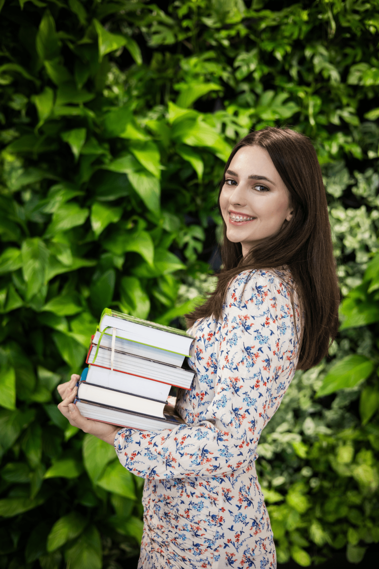 Poznaj nas | Uniwersytet WSB Merito Gdynia: Zdjęcie młodej studentki z aparatem na zębach. Ma brązowe włosy do ramion i delikatną białą sukienkę w małe kwiatki. Trzyma w ręku stertę książek. Pozuje na tle zielonej ściany z kwiatami