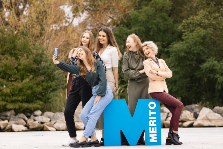 Cyfrowy uniwersytet | Uniwersytet WSB Merito Gdynia: Zdjęcie grupowe studentek, które robią sobie selfie na zewnątrz. Opierają się one o sygnet litery "m"