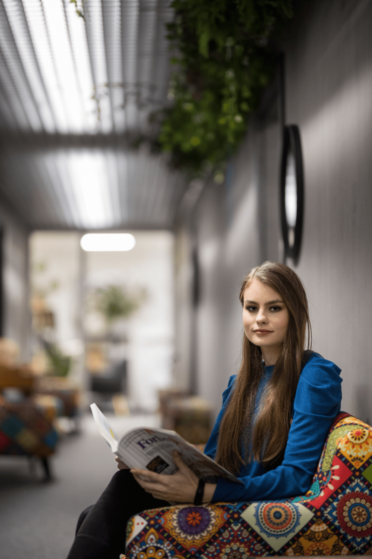 Szkoły | Uniwersytet WSB Merito Gdynia: W szarej przestrzeni w fotelu siedzi młoda studentka z gazetą na kolanach. Siedzi w patchworkowym kolorowym fotelu