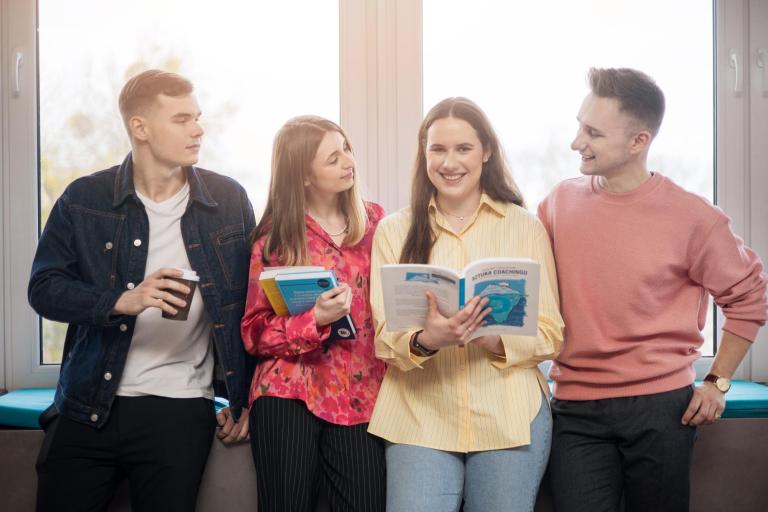 Akredytacja IACBE | Uniwersytet WSB Merito Gdynia: Zdjęcie studentów opierających się o parapet, z tyłu świeci słońce. Na zdjęciu znajduje się dwóch chłopaków i dwie dziewczyny, które zajmują centralne miejsce i trzymają książki. Wzrok wszystkich podąża za studentką, która patrzy wprost w obiektyw