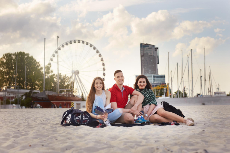 Szkoły | Uniwersytet WSB Merito Gdynia: Fotografia robiona na plaży w Gdyni, z tyłu widać wieżowiec i koło widokowe. Na plaży siedzą dwie studentki i student