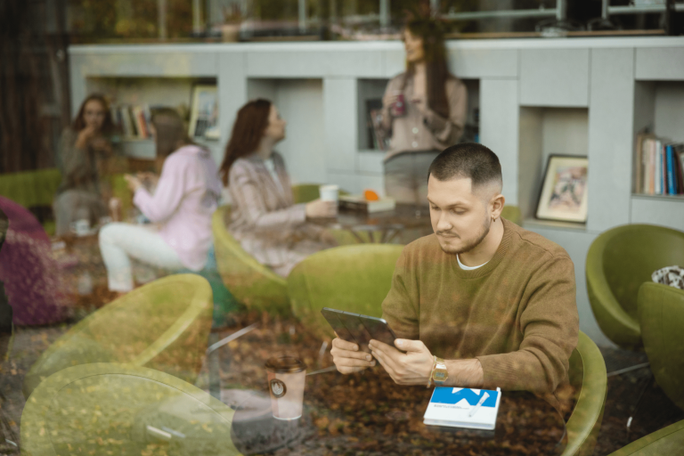 Cyfrowy uniwersytet | Uniwersytet WSB Merito Gdynia: Zdjęcie mężczyzny zza szyby, który przegląda tablet i znajduje się w kawiarni. Z tyłu widać rozmawiające kobiety. Zdjęcie jest w odcieniach beżów