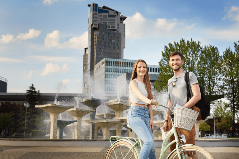Studia I stopnia w Gdyni | Uniwersytet WSB Merito Gdynia: Młodzi ludzie przy fontannie i wieżowcu w Gdyni. Dziewczyna jest na rowerze miejski, a chłopak stojący obok niej ma plecak