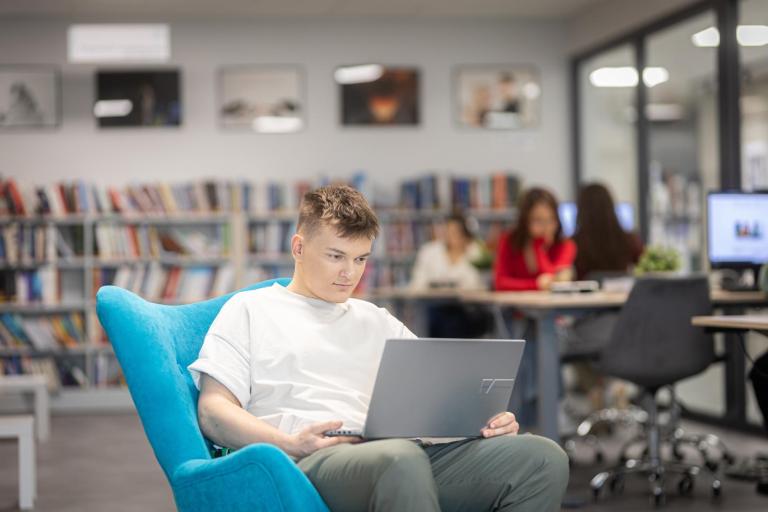 Biblioteka | Uniwersytet WSB Merito Gdynia: Student w przestrzeni biblioteki siedzi na fotelu i na kolanach trzyma laptopa