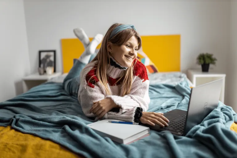Pedagogika resocjalizacyjna z profilaktyką społeczną | Studia I stopnia w Gdyni | Uniwersytet WSB Merito Gdynia: Zdjęcie studentki siedzącej na łóżku przy laptopie. Uśmiecha się, przestrzeń jest w kolorach miodowych i turkusowych