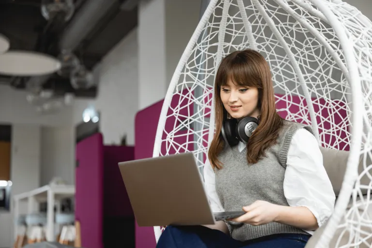 Front-end Developer | Studia I stopnia w Gdyni | Uniwersytet WSB Merito Gdynia: Zdjęcie dziewczyny siedzącej w restauracji, w ozdobnym ażurowym krześle. Przegląda ona laptopa. Ma brązowe włosy, jasne ubrania, w przestrzeni widać odcienie burgundu 