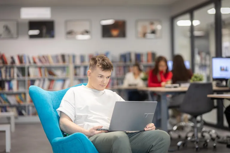 Student w przestrzeni biblioteki siedzi na fotelu i na kolanach trzyma laptopa
