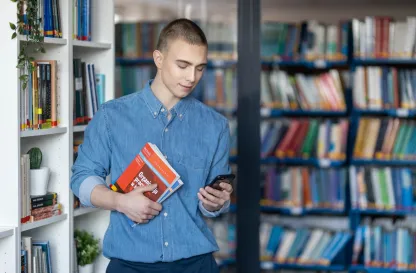 Poradniki - dzielimy się wiedzą | Uniwersytet WSB Merito: Chłopak w bibliotece trzymający telefon