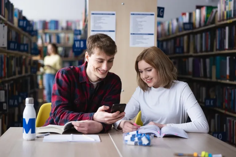 Studia związane z AI. Zastosowanie sztucznej inteligencji w praktyce | Uniwersytet WSB Merito: Dwie osoby patrzące w telefon, siedząc w bibliotece