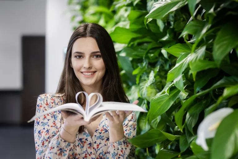Czy nadaje się na studia? Waham się, nie chcę iść na studia lub mam dość studiów: nie daję rady, nie idzie mi | Uniwersytet WSB Merito: Uśmiechnięta dziewczyna z książką, w której robi serce z kartek