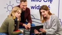 Projektowanie interfejsów graficznych | Uniwersytet WSB Merito Łódź: Troje młodych ludzi wspólnie ogląda coś na tablecie, siedząc w nowoczesnym wnętrzu z napisem „Miło Cię widzieć” w tle.