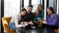Bezpieczeństwo i higiena pracy | Uniwersytet WSB Merito Łódź: Cztery młode osoby siedzą przy okrągłym stole w jasnym pomieszczeniu z dużymi oknami, skupione na wspólnej grze karcianej i uśmiechające się.
