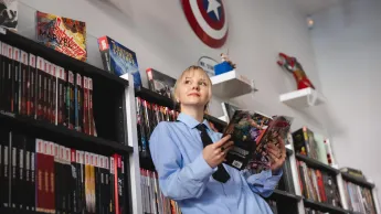 Marketing i sprzedaż | Studia I stopnia w Łodzi | Uniwersytet WSB Merito Łódź: Kobieta w niebieskiej koszuli czyta komiks, otoczona półkami pełnymi komiksów i dekoracjami Marvela.