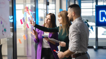 SAP S/4 HANA – Nowoczesny system ERP | Studia podyplomowe w Łodzi | Uniwersytet WSB Merito Łódź: Trzy osoby współpracują przy tablicy z kolorowymi karteczkami, omawiając pomysły i planując działania w nowoczesnym biurze.