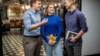 Psychologia uzależnień | Studia podyplomowe w Łodzi | Uniwersytet WSB Merito Łódź: Trójka studentów rozmawia ze sobą w przestrzeni coworkingowej, trzymając książki i uśmiechając się podczas dyskusji.