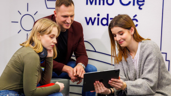 Projektowanie interfejsów graficznych | Studia podyplomowe w Łodzi | Uniwersytet WSB Merito Łódź: Troje młodych ludzi wspólnie ogląda coś na tablecie, siedząc w nowoczesnym wnętrzu z napisem „Miło Cię widzieć” w tle.