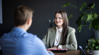 Psychologia sądowo - penitencjarna | Studia jednolite magisterskie w Łodzi | Uniwersytet WSB Merito Łódź: Kobieta w okularach i eleganckim stroju uśmiecha się podczas rozmowy z mężczyzną w jasnoniebieskiej koszuli przy biurku w nowoczesnym biurze.
