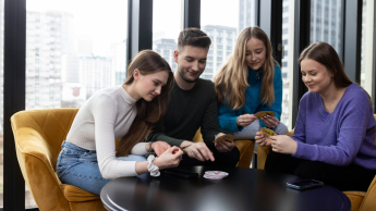 Bezpieczeństwo i higiena pracy | Studia podyplomowe w Łodzi | Uniwersytet WSB Merito Łódź: Cztery młode osoby siedzą przy okrągłym stole w jasnym pomieszczeniu z dużymi oknami, skupione na wspólnej grze karcianej i uśmiechające się.