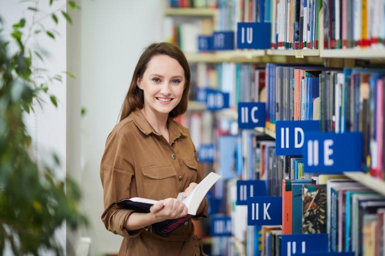 Biblioteka | Uniwersytet WSB Merito Łódź: Kobieta w bibliotece trzyma książkę i uśmiecha się