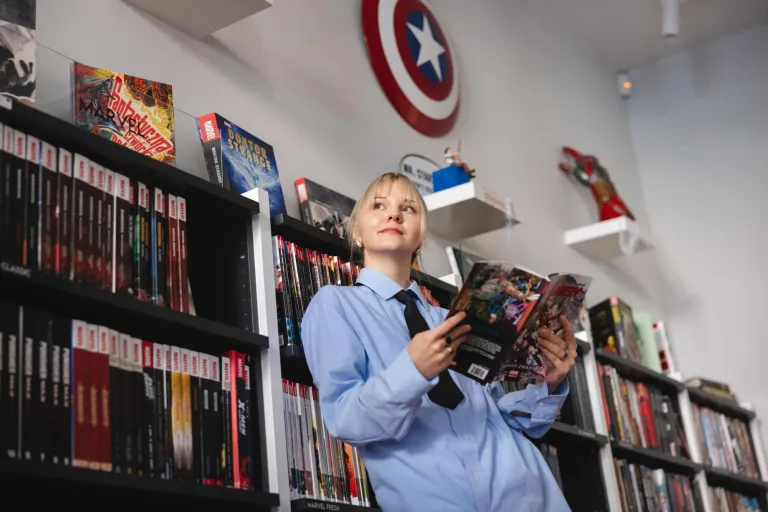 Stypendia i zniżki | Uniwersytet WSB Merito Łódź: Kobieta w niebieskiej koszuli czyta komiks, otoczona półkami pełnymi komiksów i dekoracjami Marvela.