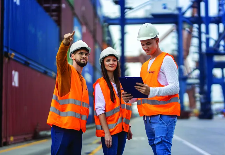 Logistyka | Studia I stopnia w Łodzi | Uniwersytet WSB Merito Łódź: Trójka młodych ludzi w kaskach i kamizelkach odblaskowych omawia dokumentację na tle kontenerów w porcie, zdobywając praktyczne doświadczenie w logistyce.
