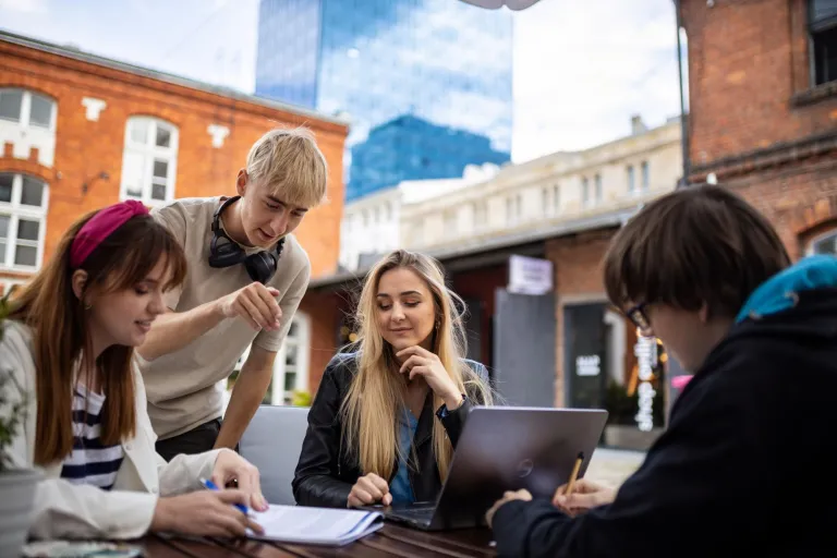 Logistyka służb mundurowych | Studia I stopnia w Łodzi | Uniwersytet WSB Merito Łódź: Grupa młodych osób pracuje wspólnie nad projektem na świeżym powietrzu, korzystając z laptopa i notatek w miejskim otoczeniu.