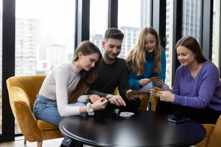 Prawo nowoczesnych technologii i cyberbezpieczeństwo | Studia I stopnia w Łodzi | Uniwersytet WSB Merito Łódź: Cztery młode osoby siedzą przy okrągłym stole w jasnym pomieszczeniu z dużymi oknami, skupione na wspólnej grze karcianej i uśmiechające się.