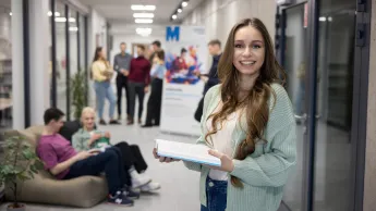 Psychodietetyka | Studia I stopnia w Opolu | Uniwersytet WSB Merito Opole: Główna grafika: Opole - psychodietetyka - uśmiechnięta studentka stojąca na korytarzu, trzyma książkę