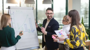 Planowanie produkcji – nowoczesne   narzędzia: SAP S/4HANA, Siemens Opcenter APS | Studia podyplomowe w Opolu | Uniwersytet WSB Merito Opole: Główna grafika: Opole - studia podyplomowe - Planowanie produkcji – nowoczesne   narzędzia: SAP S/4HANA, Siemens Opcenter APS - Grupa osób rozpisująca wykresy przy białej tablicy