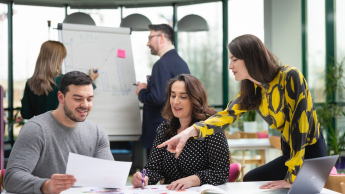 Psychologia reklamy i komunikacji | Studia podyplomowe w Opolu | Uniwersytet WSB Merito Opole: Główna grafika: Opole - studia podyplomowe - Psychologia reklamy i komunikacji - Grupa osób pracująca nad projektem, w tle dwie osoby piszące na białej tablicy