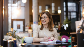 Energetyka wiatrowa | Uniwersytet WSB Merito Opole: Główna grafika: Opole - studia podyplomowe - Energetyka wiatrowa (Onshore & Offshore) - Kobieta siedząca w kawiarni, ma przed są otwarty notatnik