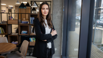 Negocjacje w biznesie | Studia podyplomowe w Opolu | Uniwersytet WSB Merito Opole: Główna grafika: Opole - studia podyplomowe - Negocjacje w biznesie - Kobieta stojąca w kawiarni, za nią regał na książki