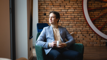 Sztuczna inteligencja w marketingu cyfrowym | Uniwersytet WSB Merito Opole: Główna grafika: Opole - studia podyplomowe - Sztuczna inteligencja w marketingu cyfrowym - Mężczyzna siedzący w zielonym fotelu, w rękach trzyma kubek kawy