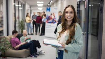 Służba cywilna w administracji | Studia I stopnia w Opolu | Uniwersytet WSB Merito Opole: Główna grafika: Opole - studia I stopnia - specjalność służba cywilna w administracji - Dziewczyna stojąca na korytarzy uczelni, trzyma książkę