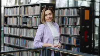 Mediacje sądowe i pozasądowe     | Studia podyplomowe w Opolu | Uniwersytet WSB Merito Opole: główna grafika: Opole - studia podyplomowe - mediacje sądowe i pozasadowe - kobieta w jasnej marynarce stoi przy barierce a za sobą ma regały z książkami