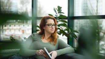 Główna grafika: Opole - studia podyplomowe - Terapia uzależnień - uczestniczka studiów podyplomowych siedzi na kanapie z książką w rękach i spogląda za okno