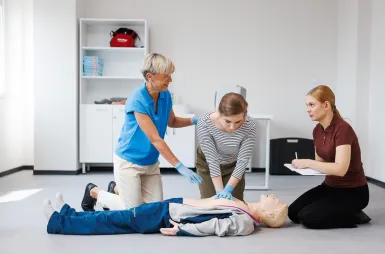 Laboratoria i pracownie | Uniwersytet WSB Merito Opole: Pracownia pierwszej pomocy: dwie studentki uczą się od pani profesor o tym, jak poprawnie przeprowadzać resuscytację na manekinie przedstawiającym dorosłego mężczyznę