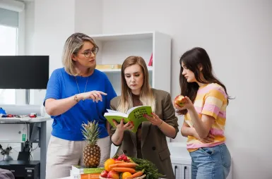 Laboratoria i pracownie | Uniwersytet WSB Merito Opole: Laboratorium dietetyczne, trzy studentki czytają książkę, jedna z nich je jabłko
