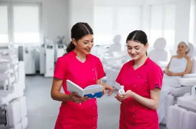 Laboratoria i pracownie | Uniwersytet WSB Merito Opole: Laboratorium kosmetologiczne, dwie studentki w różowych fartuchach rozmawiają, w tle białe fotele