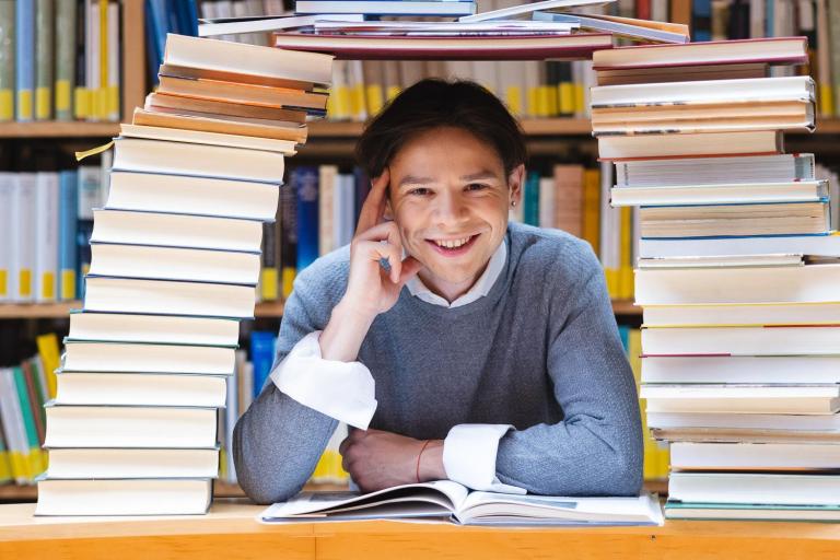 Biblioteka | Uniwersytet WSB Merito Opole: Chłopak w bibliotece. Siedzi pod mostem zbudowanym z książek.