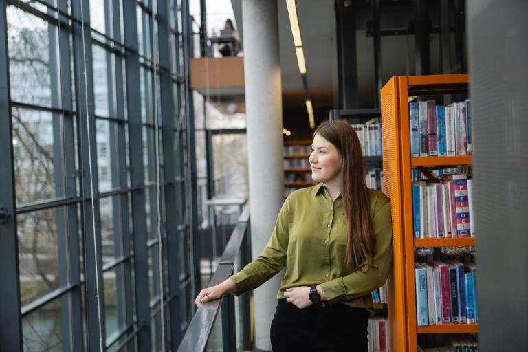 Dział Studiów Podyplomowych | Uniwersytet WSB Merito Opole: W bibliotece stoi kobieta w zielonej koszuli i wygląda za okno